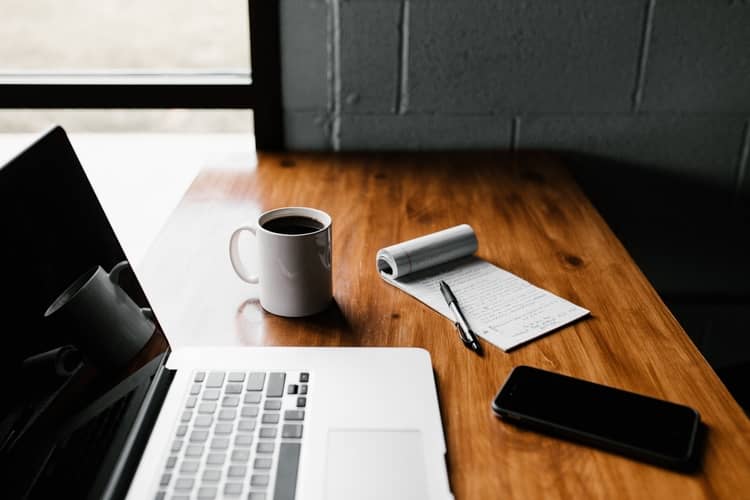 a desk with a laptop, coffee mug, notepad and pen and a smartphone