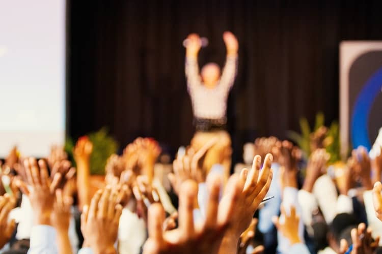 an out of focus image of a public speaker and their audience with raised hands.